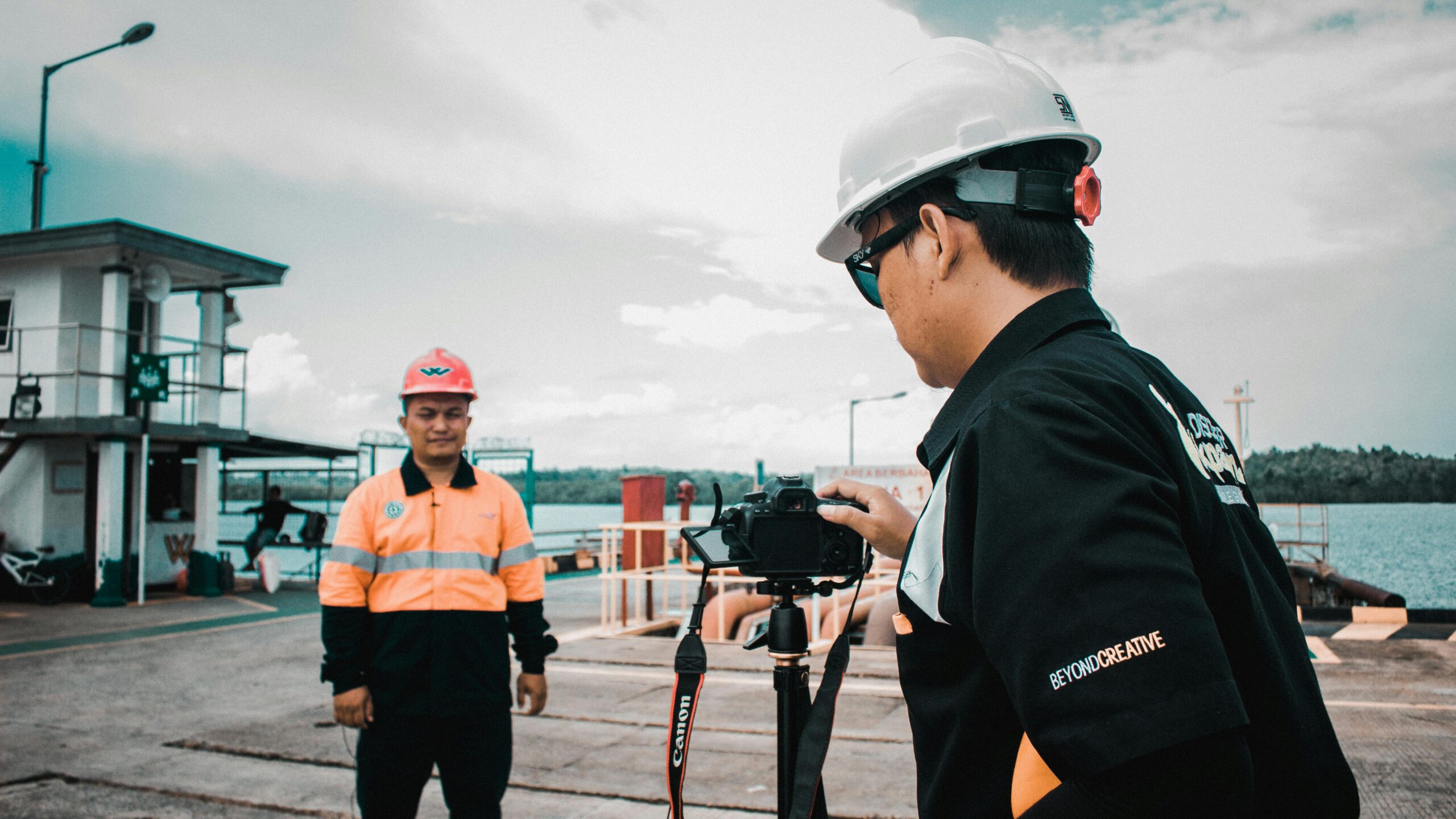 Two construction workers in safety gear filming at a waterfront location.